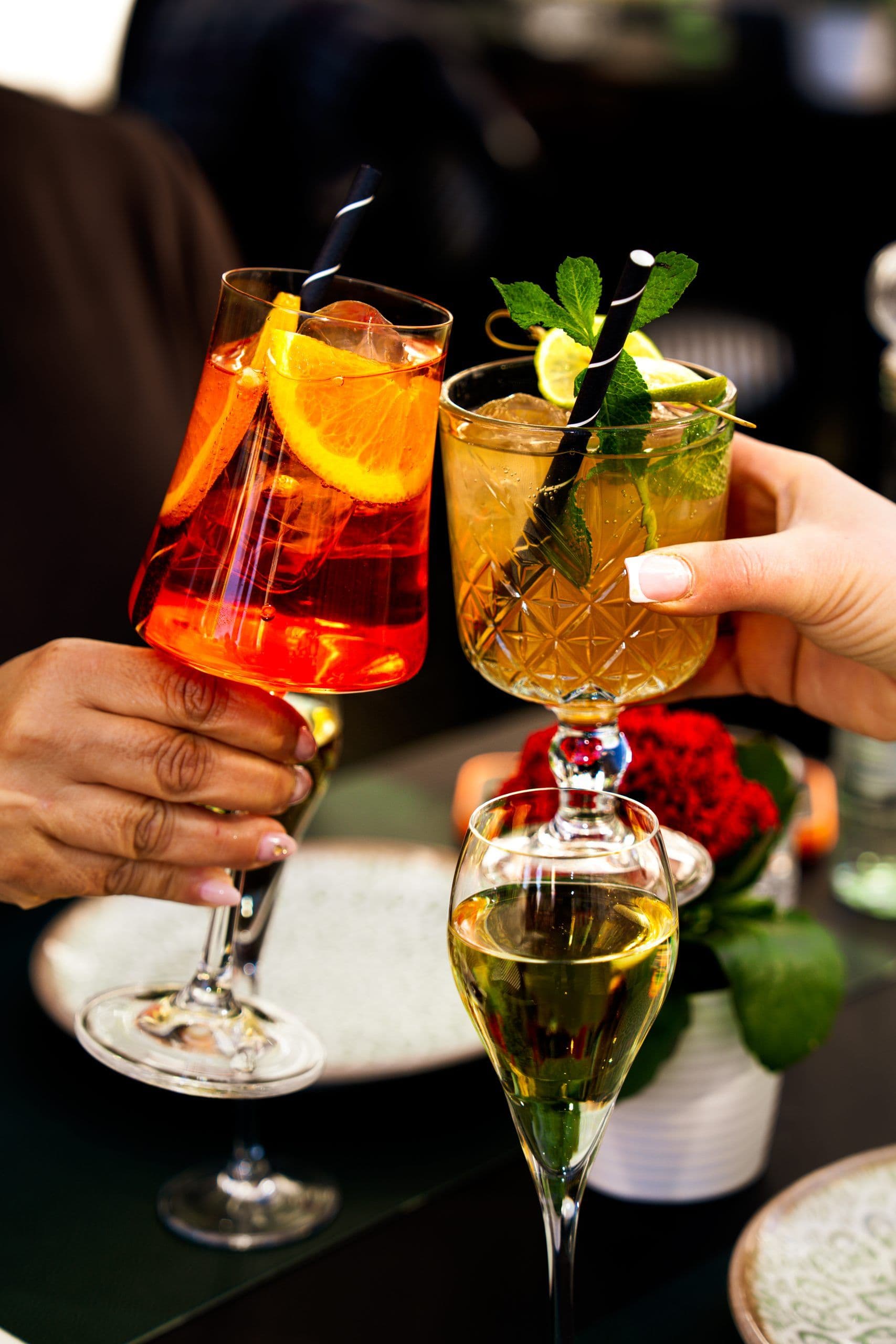 Zwei Hände stossen mit einem orangefarbenen Aperitif und einem Longdrink mit Minze und Limette an.
