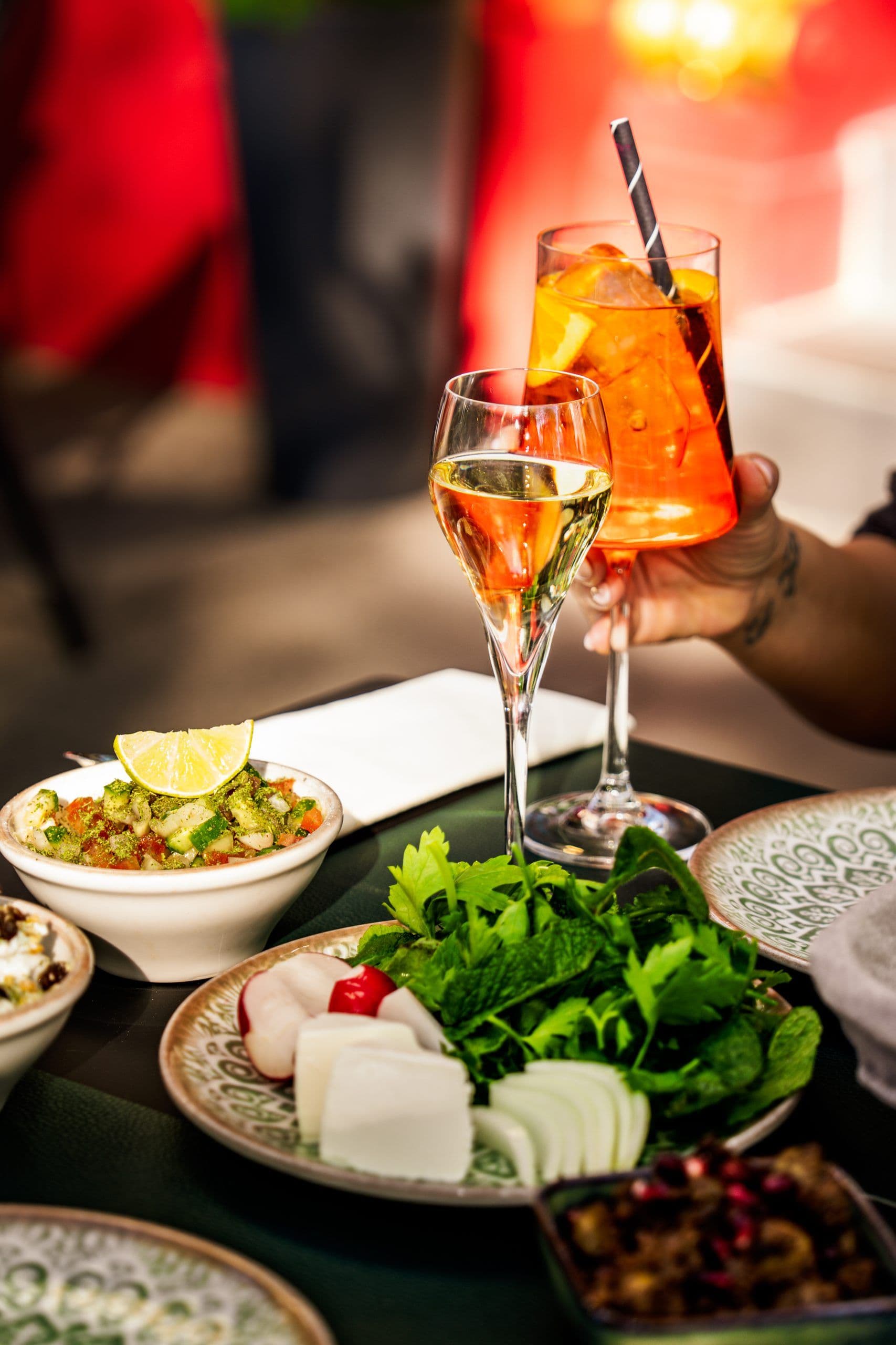 Glas Weißwein und oranger Spritz neben Mezze mit Kräutern, weißem Käse und Radieschen.