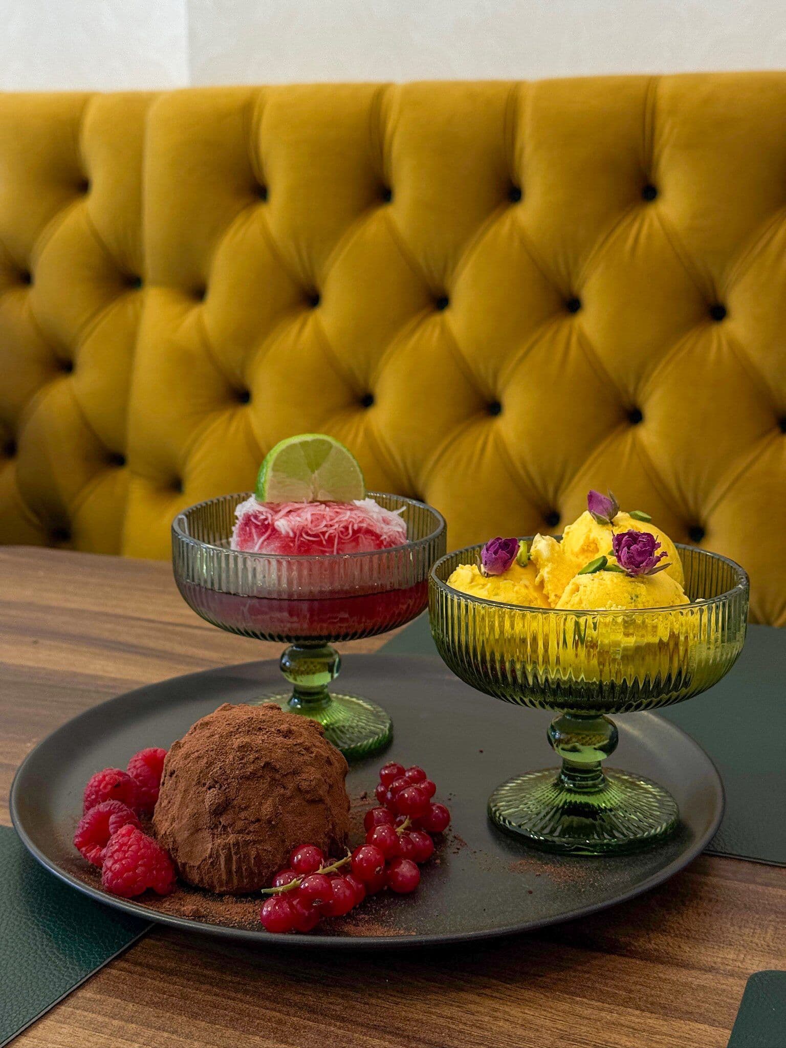 Drei Desserts vor gelber Samtbank: Safraneis mit Pistazien und Rosenknospen, rosa Sorbet mit Limette und Kakao-Dessert mit Beeren.