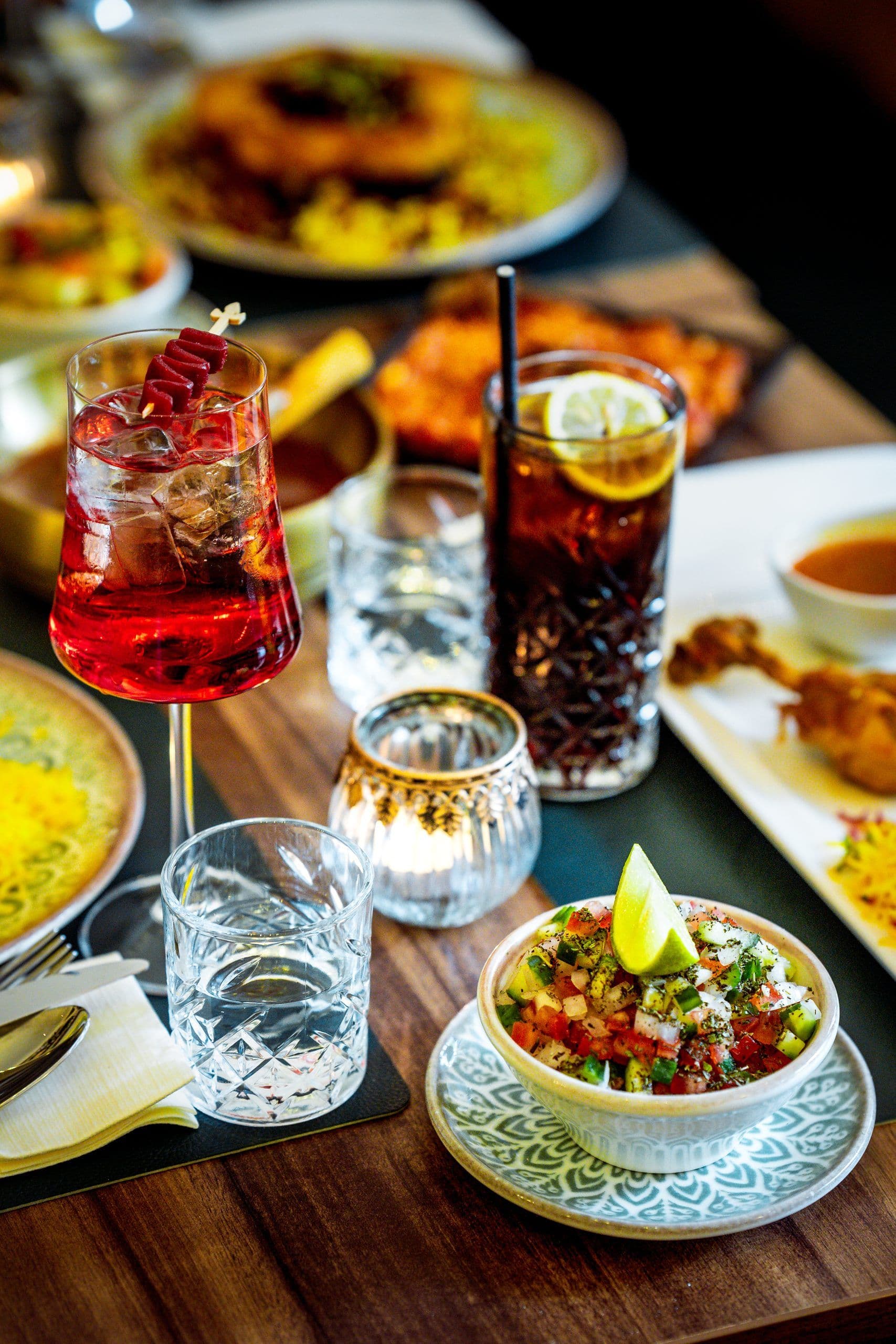 Roter Aperitif im Stielglas, dunkler Longdrink und Schale mit gewürfeltem Salat auf gedeckter Tafel.