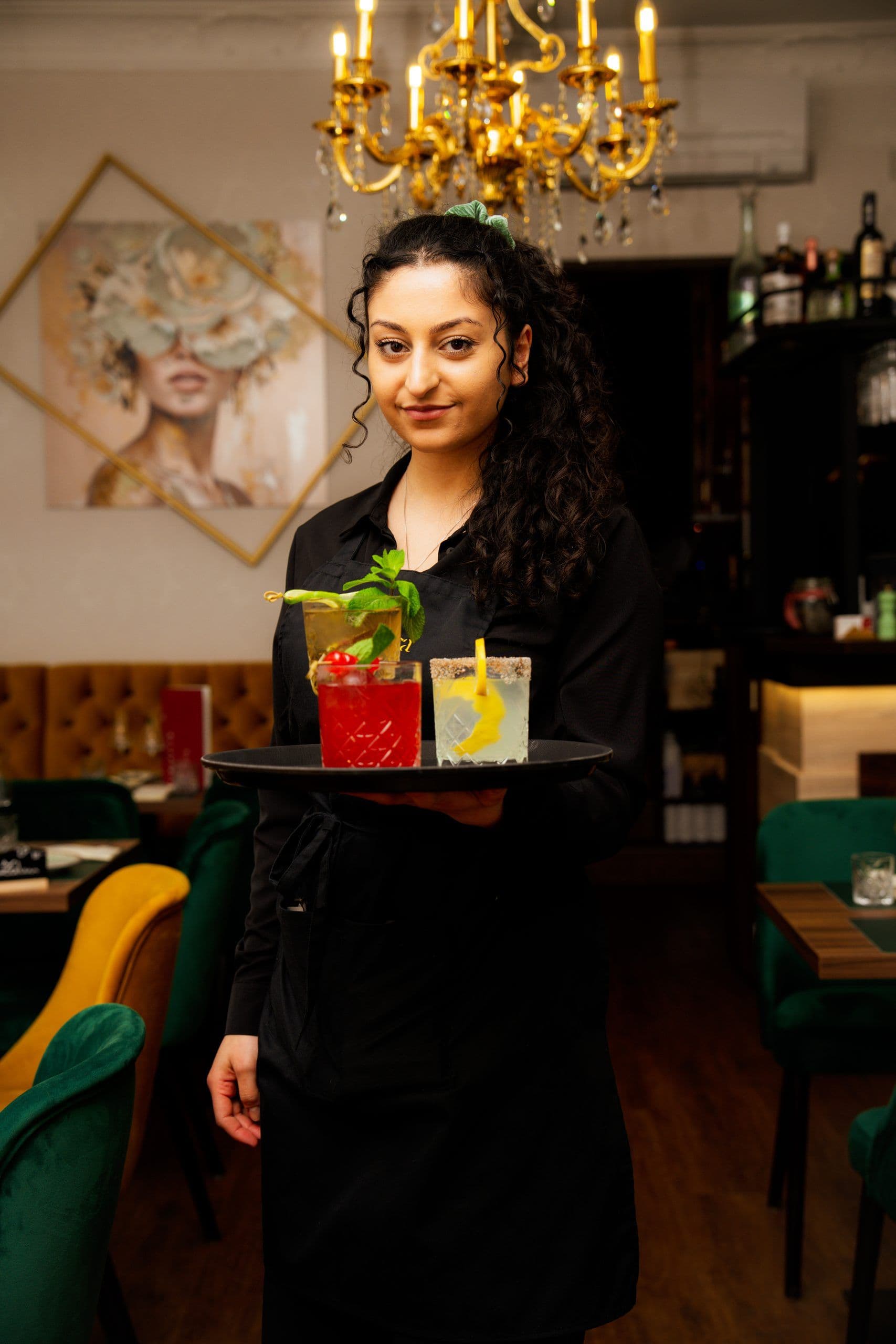 Mitarbeiterin im Service hält ein Tablett mit drei garnierten Cocktails im Gastraum unter dem Kronleuchter.